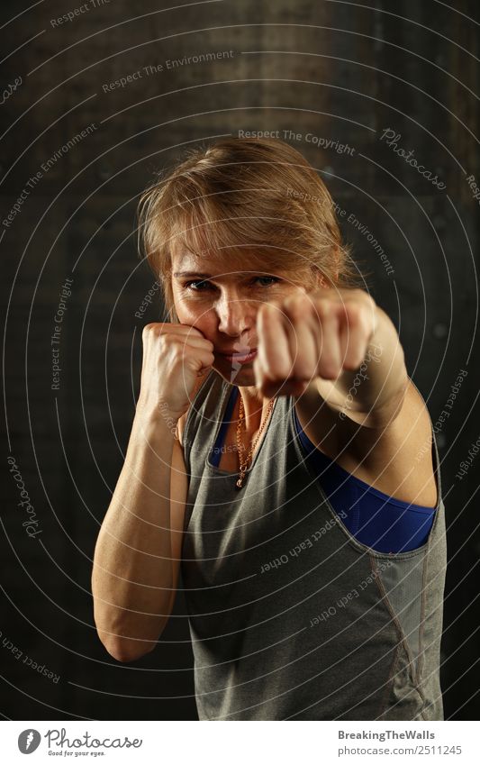 Close up front portrait of one young middle age athletic woman in sportswear in gym over dark background, standing in boxing stance with hands and fists, looking at camera