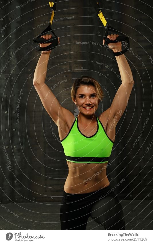 One young athletic woman at crossfit training, exercising with trx suspension fitness straps over dark background, front view, looking at camera Lifestyle