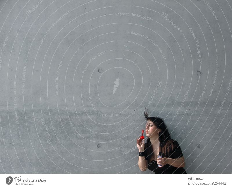 Woman blowing soap bubbles with abandon in front of grey wall, neutral background Adults 1 Human being Wall (barrier) Wall (building) Brunette Playing Gray Ease