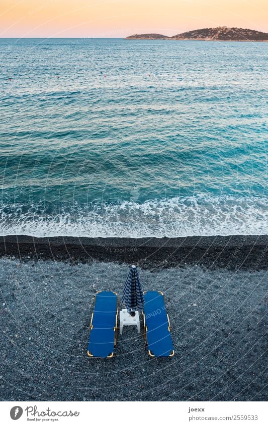 Two blue beach chairs with parasol and small tables on the dark beach with turquoise sea in front of sunset from above Lie Blue Sunshade Beach Black