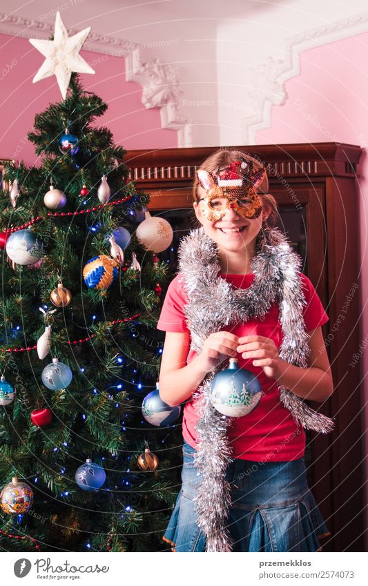 Young girl decorating Christmas tree, holding big Christmas ball. Teenage blonde girl wearing blue jeans skirt and pink blouse and tights. Girl has a funny reindeer mask on her face. Young girl celebrating Christmas at home