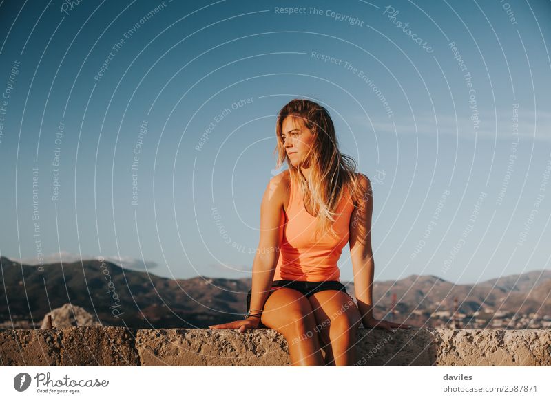 Beautiful woman with sports clothes, sitting on a concrete wall outdoors at sunset. Lifestyle Sun Mountain Sports Fitness Sports Training Human being Feminine