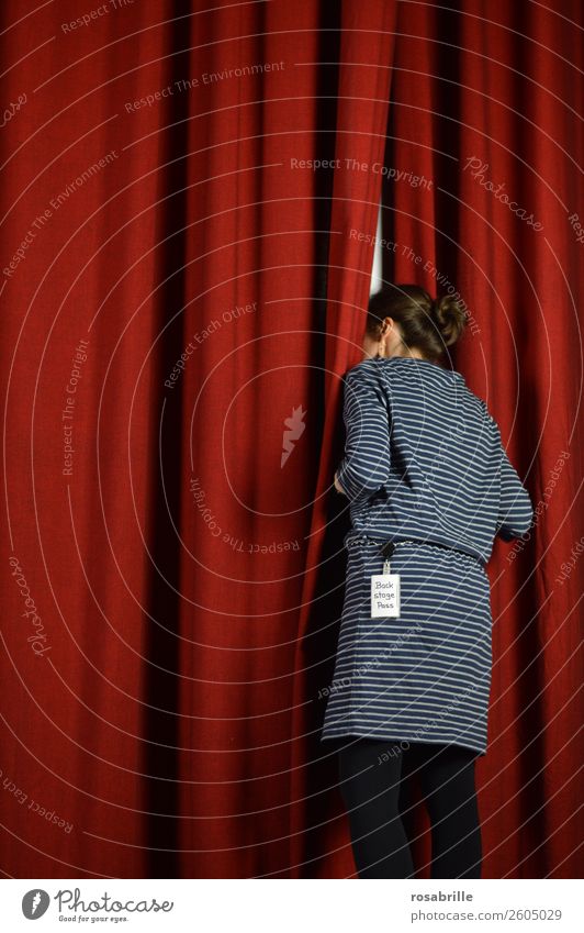 young woman in a blue dress dares to take a look behind the scenes and curiously looks behind a red theatre curtain as a symbol for background information, insights or announcements