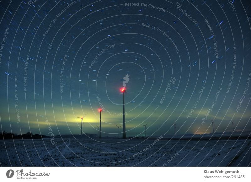 Wind turbines stand under a starry sky on a snow-covered field. The light of a distant city is reflected by clouds. Energy industry Renewable energy