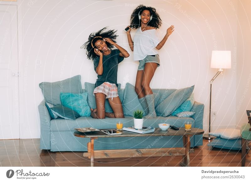 Happy young two black women sitting and standing up in the couch on the computer and mobile phone with headphones dancing Woman Computer Cellphone Mobile