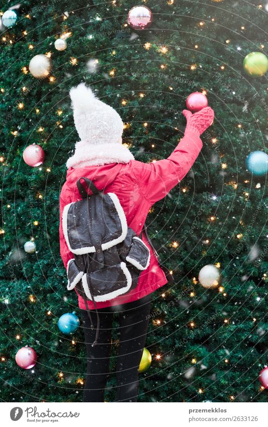 Young girl standing at front of big Christmas tree decorated with colorful balls. Teenager girl is wearing warm clothes, scarf and wool cap on a cold winter day