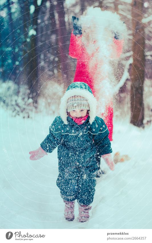 Mother is playing with her little daughter outdoors on wintery day. Woman is throwing snow on her child. Family spending time together enjoying wintertime. Woman is wearing red coat and wool cap, toddler is wearing dark blue snowsuit