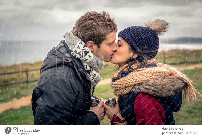 Young couple kissing and holding cups of hot drink outdoors Beverage Coffee Lifestyle Adventure Ocean Winter Mountain Woman Adults Man Couple Hand Nature Sky