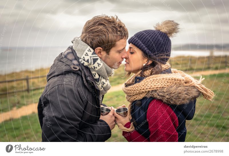 Young couple kissing and holding cups of hot drink outdoors Beverage Coffee Lifestyle Happy Ocean Winter Mountain Woman Adults Man Couple Hand Nature Sky Clouds