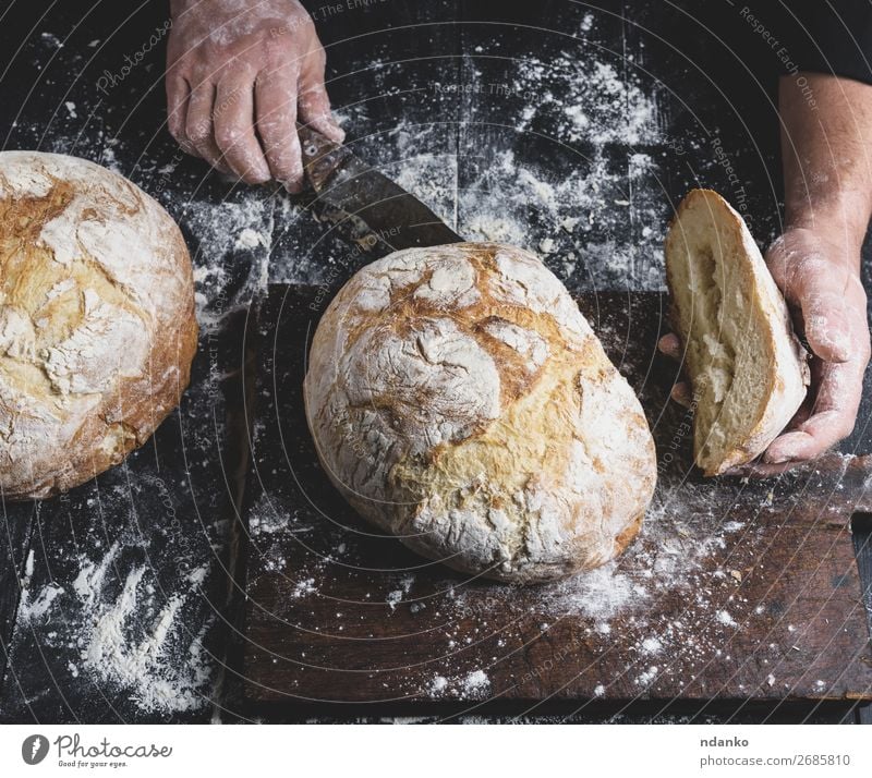 man cuts baked round bread on a brown wooden board Bread Nutrition Table Kitchen Human being Hand Fingers Wood Make Dark Fresh Brown Black White Tradition whole