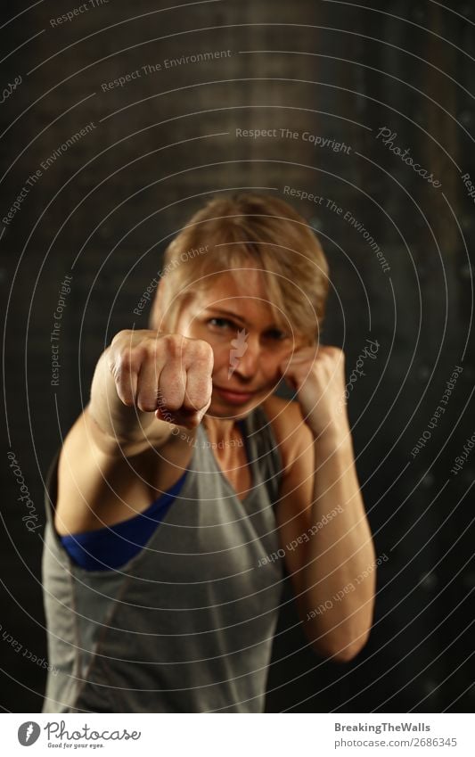 Close up front portrait of one young middle age athletic woman in sportswear in gym over dark background, standing in boxing stance with hands and fists, looking at camera