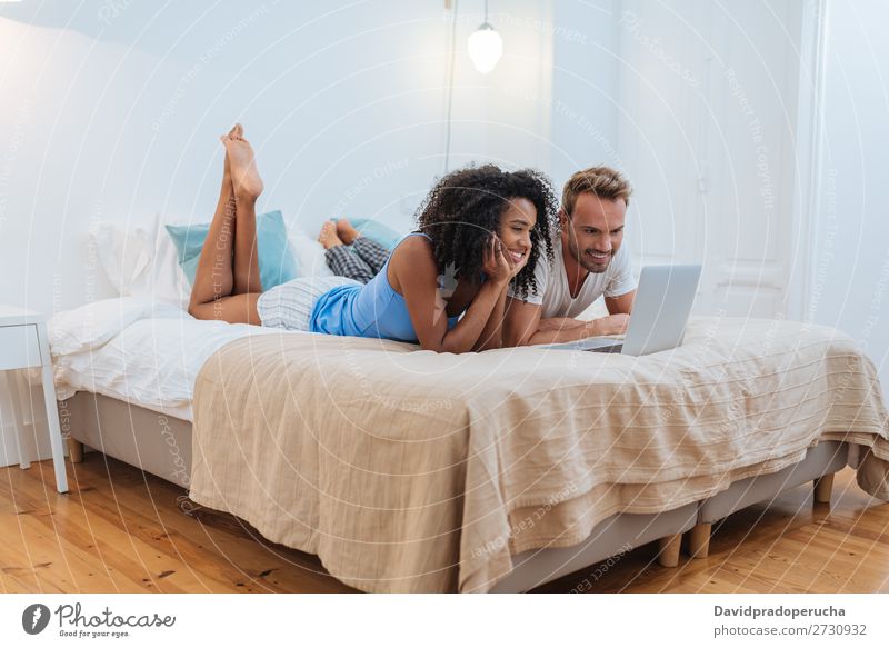 Happy young couple relaxed at home lying down in bed on the computer interracial Relationship Couple Youth (Young adults) Bed Bedroom Technology Computer