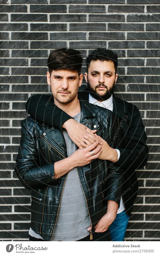 Gay couple hugging near brick wall gay embrace looking at camera happy standing posing homosexual pair male love two together lifestyle relationship young men