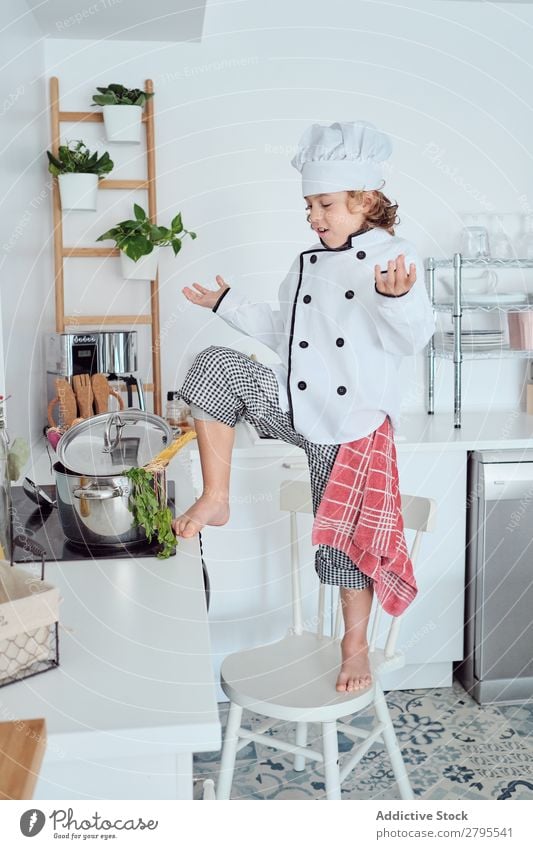 Boy in cook hat with upped hand near pot on chair in kitchen Cook Boy (child) Pot Hand Kitchen Chair chef Child Vegetable Hat Cooking Modern Funny Home Light