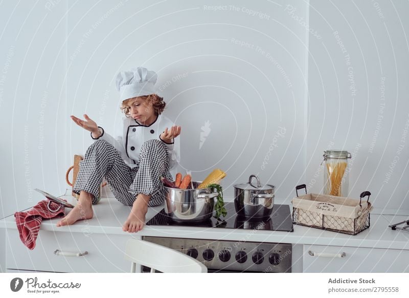 Boy in cook hat sitting near pots on electric fryer in kitchen Cook Boy (child) Pot Kitchen chef Child Vegetable Hat Stove & Oven Cooking Modern Funny Home