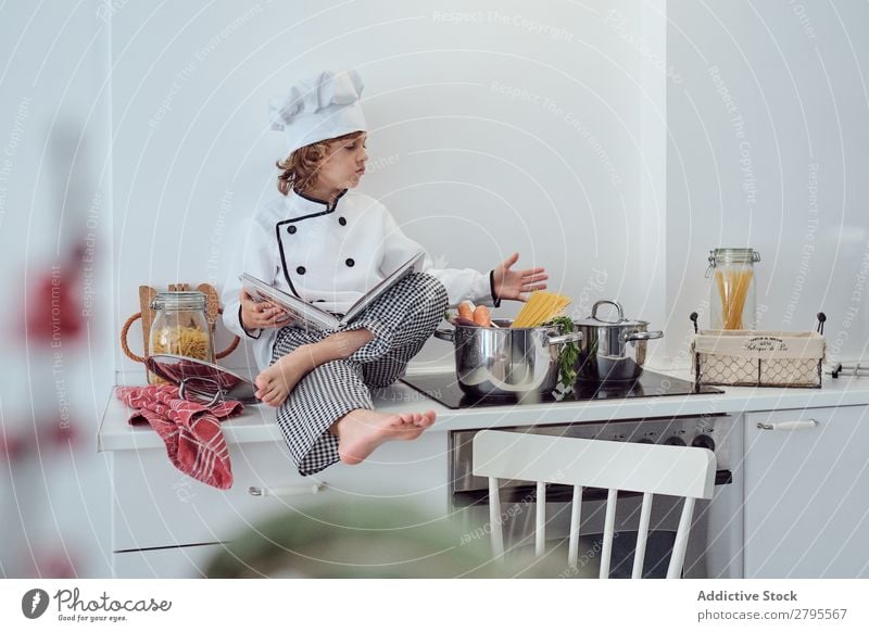 Boy in cook hat with book sitting near pots on electric fryer in kitchen Cook Boy (child) Book Pot Kitchen chef Child Vegetable Hat Volume Reading Stove & Oven