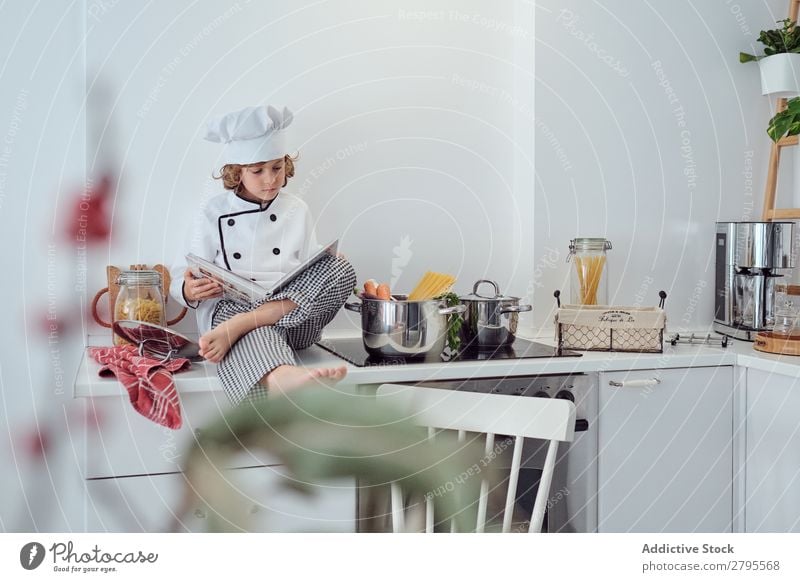 Boy in cook hat with book sitting near pots on electric fryer in kitchen Cook Boy (child) Book Pot Kitchen chef Child Vegetable Hat Volume Reading Stove & Oven