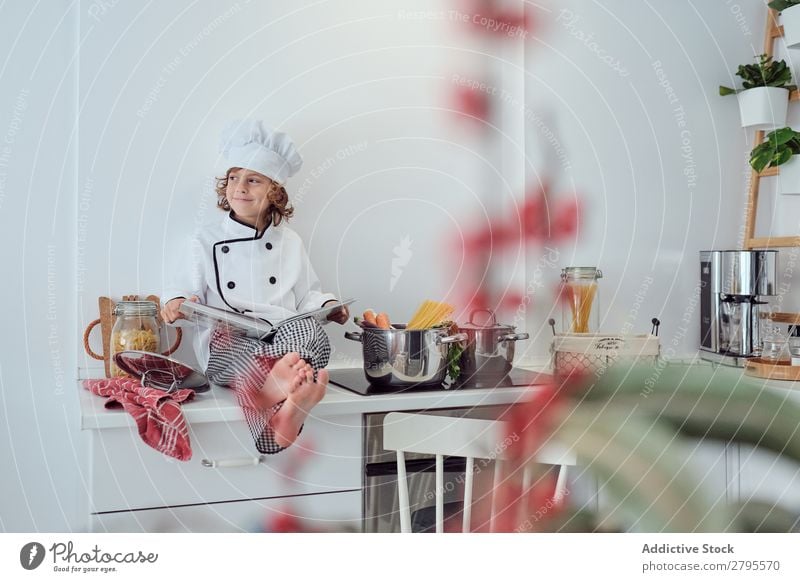 Boy in cook hat with book sitting near pots on electric fryer in kitchen Cook Boy (child) Book Pot Kitchen chef Child Vegetable Hat Volume Reading Stove & Oven