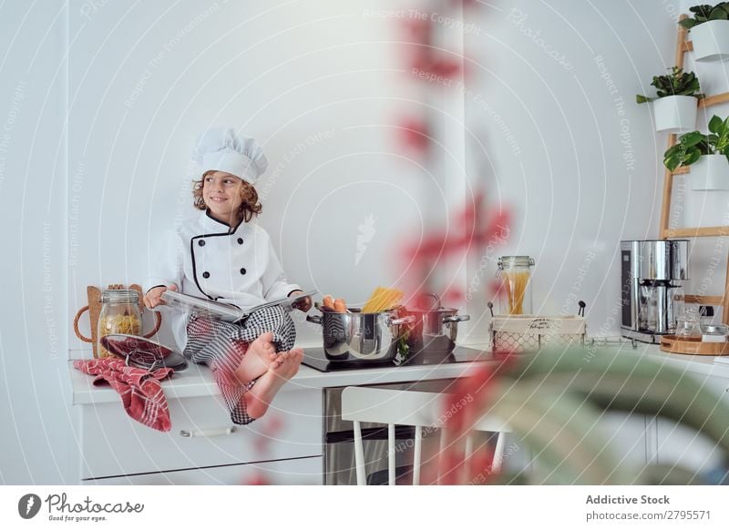 Boy in cook hat with book sitting near pots on electric fryer in kitchen Cook Boy (child) Book Pot Kitchen chef Child Vegetable Hat Volume Reading Stove & Oven