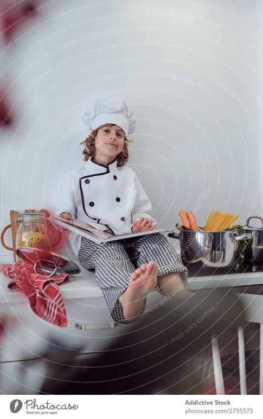 Boy in cook hat with book sitting near pots on electric fryer in kitchen Cook Boy (child) Book Pot Kitchen chef Child Vegetable Hat Volume Reading Stove & Oven