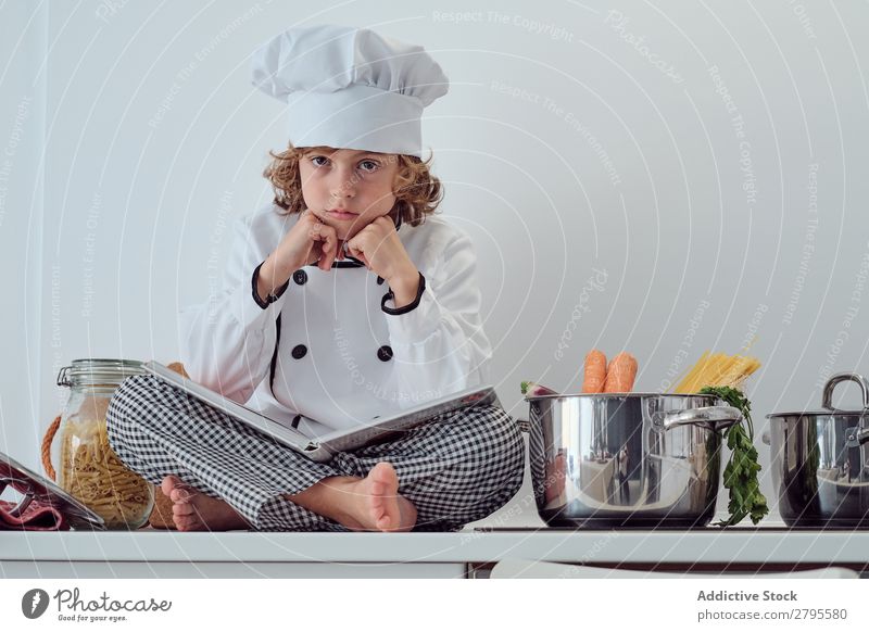 Boy in cook hat with book sitting near pots on electric fryer in kitchen Cook Boy (child) Book Pot Kitchen chef Child Vegetable Hat Volume Reading Stove & Oven