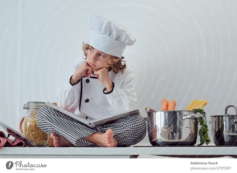 Boy in cook hat with book sitting near pots on electric fryer in kitchen Cook Boy (child) Book Pot Kitchen chef Child Vegetable Hat Volume Reading Stove & Oven