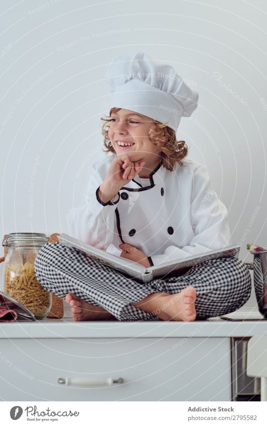 Boy in cook hat with book sitting near pots on electric fryer in kitchen Cook Boy (child) Book Pot Kitchen chef Child Vegetable Hat Volume Reading Stove & Oven