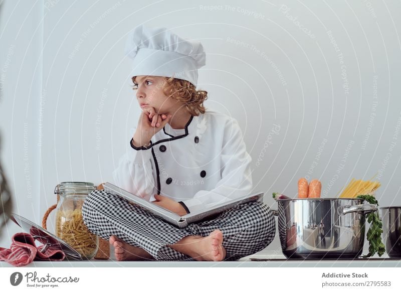 Boy in cook hat with book sitting near pots on electric fryer in kitchen Cook Boy (child) Book Pot Kitchen chef Child Vegetable Hat Volume Reading Stove & Oven