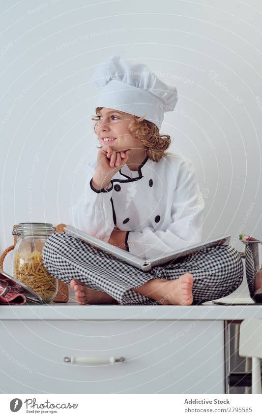 Boy in cook hat with book sitting near pots on electric fryer in kitchen Cook Boy (child) Book Pot Kitchen chef Child Vegetable Hat Volume Reading Stove & Oven