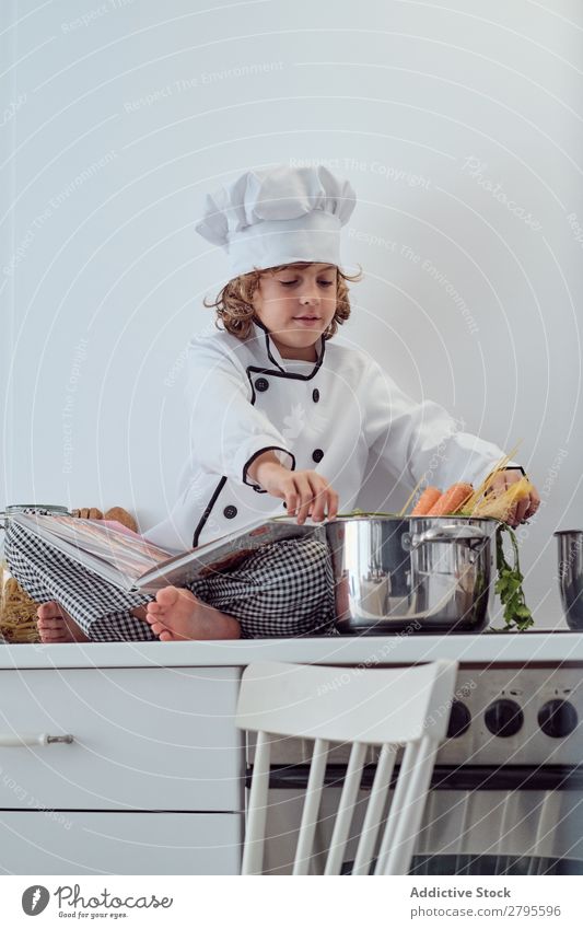 Boy in cook hat with book sitting near pots on electric fryer in kitchen Cook Boy (child) Book Pot Kitchen chef Child Vegetable Hat Volume Reading Stove & Oven