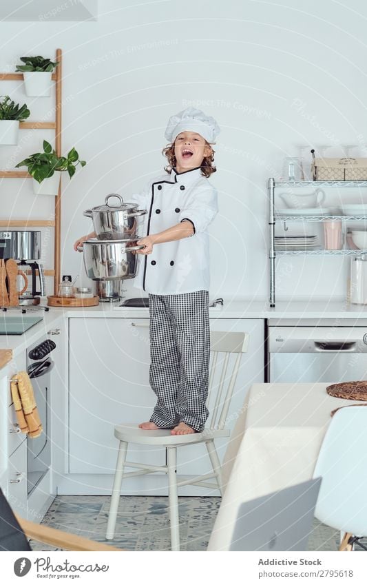 Smiling boy in cook hat holding pots on chair in kitchen Cook Boy (child) Pot Kitchen Chair chef Child Hat Cooking Modern Home Happy Light preparing Happiness