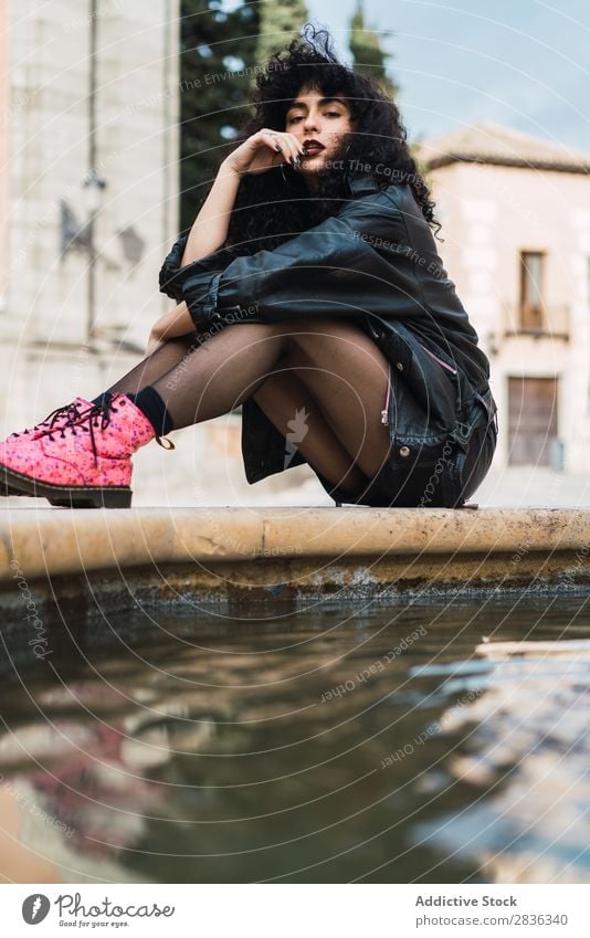 Young woman posing at fountain Woman Attractive City fashionable Curly Brunette Jacket Fountain Water Looking into the camera Fashion Youth (Young adults)