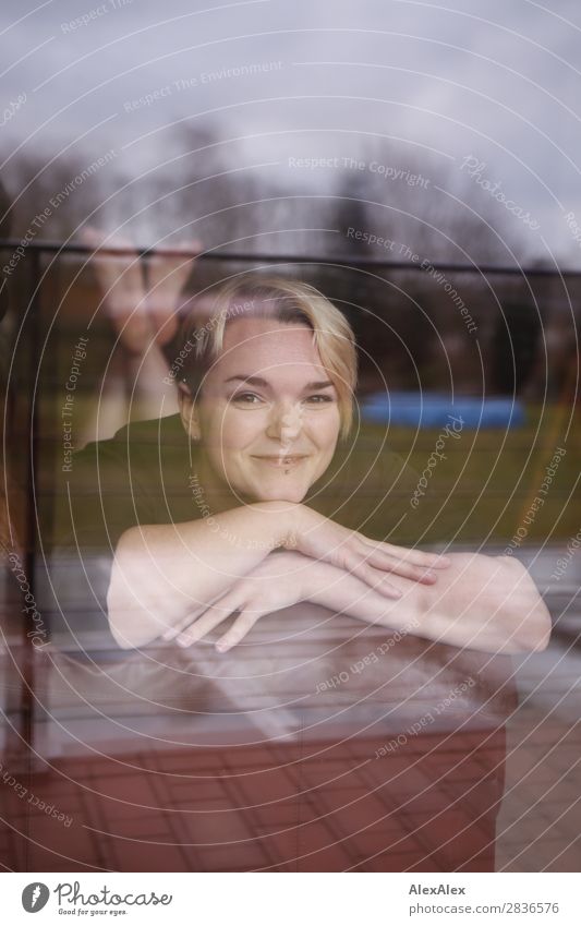 Young woman behind window lies on couch and smiles at camera Joy pretty Life Well-being Flat (apartment) Room Window Youth (Young adults) 18 - 30 years Adults