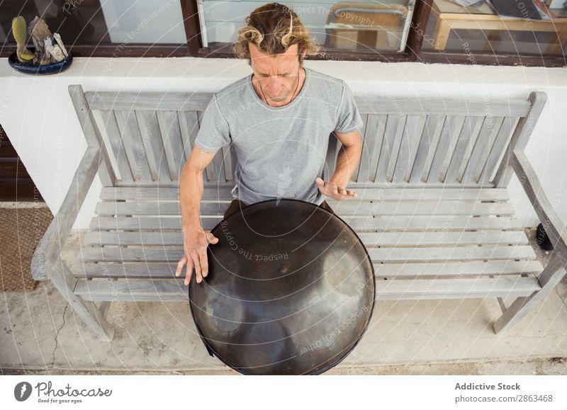 Blond man sitting on bench with big hand drum Man Drum handpan Bench Sit Hand Barefoot T-shirt Seat Blonde Window Door Building Music Guy instrument Playing