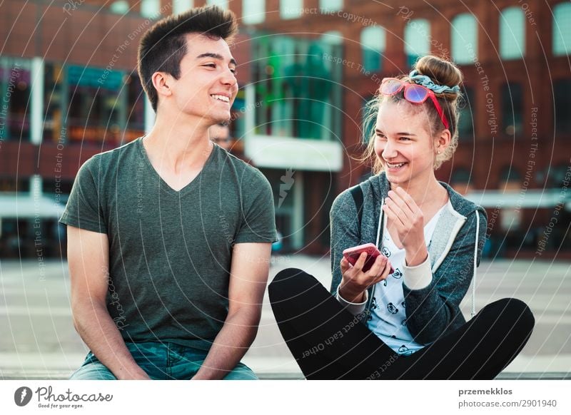 Couple of friends, teenage girl and boy, having fun together with smartphones, sitting in center of town, spending time together Lifestyle Summer To talk