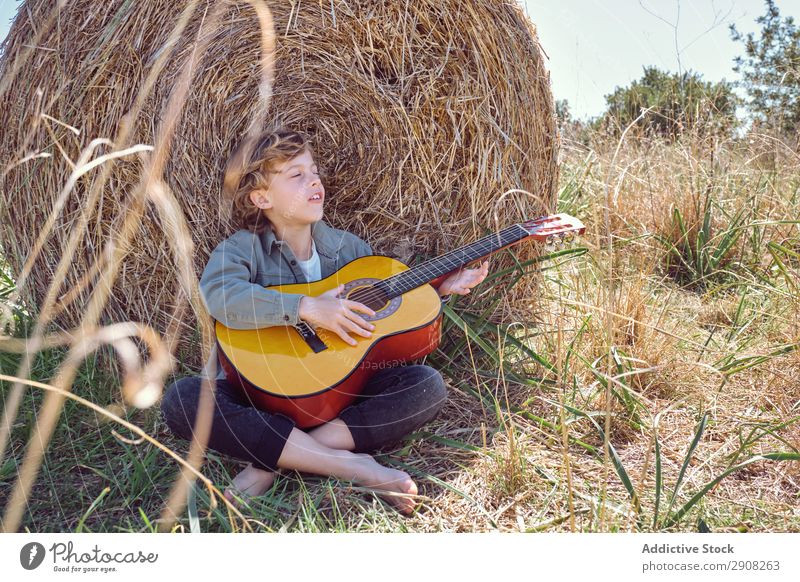 Boy playing guitar and singing near roll of hay Boy (child) Guitar Playing Music Field Hay Roll Closed eyes Landscape Child Grass Dry Song Acoustic instrument