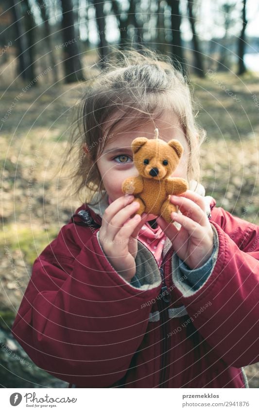 Little smilling happy girl playing with her little teddy bear toy in a park on sunny spring day. Child holding teddy at front of face looking at camera wearing red jacket