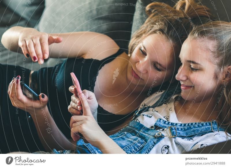 Young women using mobile phones watching music clip, texting, messaging. Teenagers using the smartphones, sitting on sofa at home. Using technology devices. Girls wearing summer clothing