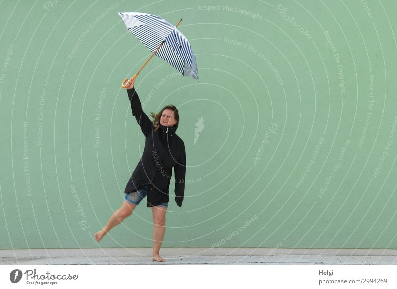 Woman with dark jacket and short jeans stands barefoot on one leg in front of a light green wall and holds up an umbrella Human being Feminine Adults 1