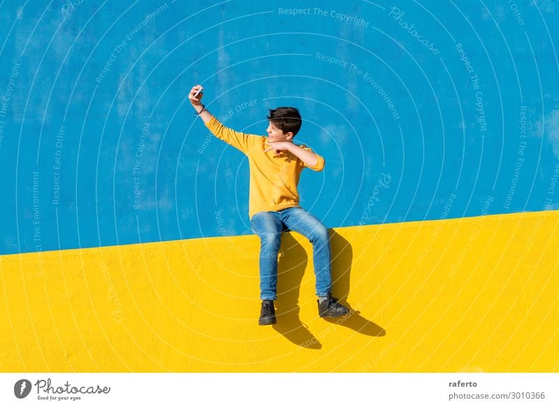 Front view of a young boy wearing casual clothes sitting on a yellow fence against a blue wall while using a mobile phone Lifestyle Summer Telephone Cellphone