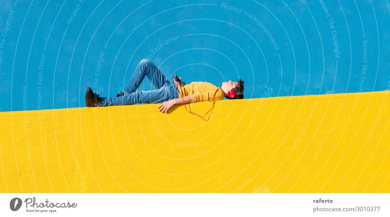 Front view of a young boy wearing casual clothes lying on a yellow fence against a blue wall while using a mobile phone to listening music Lifestyle Music