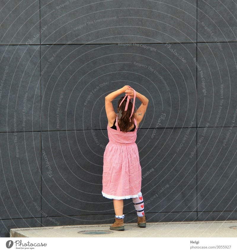 Rear view of a woman with long brunette hair, pink dress and patterned stockings standing with raised arms in front of a dark grey wall Human being Feminine