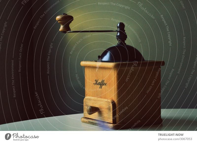 Coffee grinder for manual operation made of solid, natural coloured wood with black metal filler cap stands at the edge of a round white table in front of a grey wall in the pale morning light