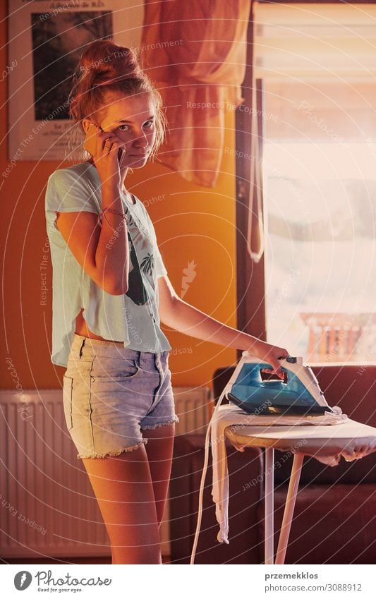 Young woman is ironing her clothes and talking on a smartphone simultaneously, standing by a ironing board in a room at home. Routine housekeeping task at home. Candid people, real moments, authentic situations