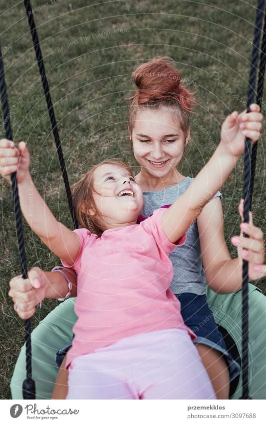 Teenage girl playing with her younger sister in a home playground in a backyard. Happy smiling sisters having fun on a swing together on summer day. Real people, authentic situations