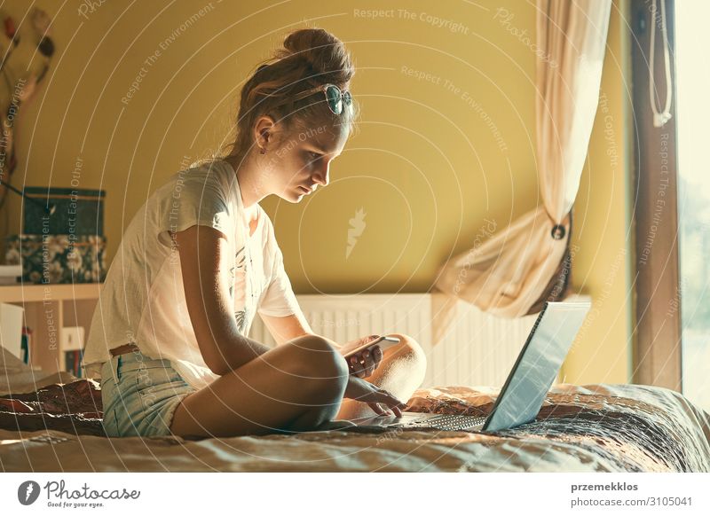 Young woman using portable computer and mobile phone, sitting on a bed, learning online at home. Candid people, real moments, authentic situations School Study