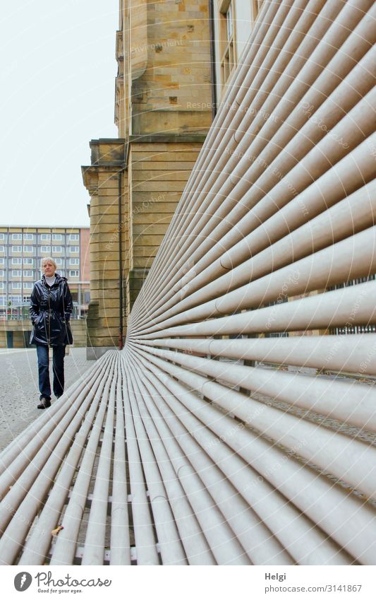 Perspective shot of a long bench in front of a building and a walking woman with a rain jacket and jeans Human being Feminine Woman Adults Female senior 1