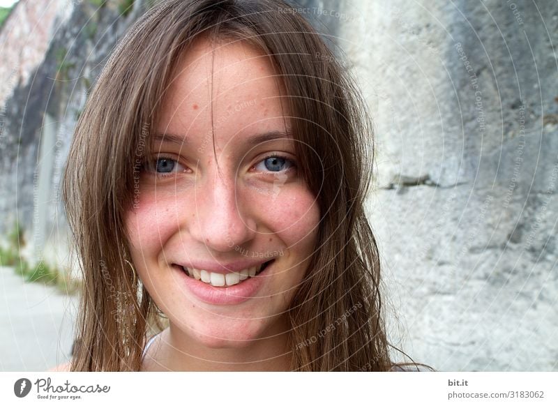 Pretty brunette, long-haired young woman, teenager, teenager smiles naturally into the camera, in front of a grey wall on vacation. Happy girl with blue eyes, in front of a rock by the sea, on the beach during the summer holidays, looking forward to holidays and summer fun.