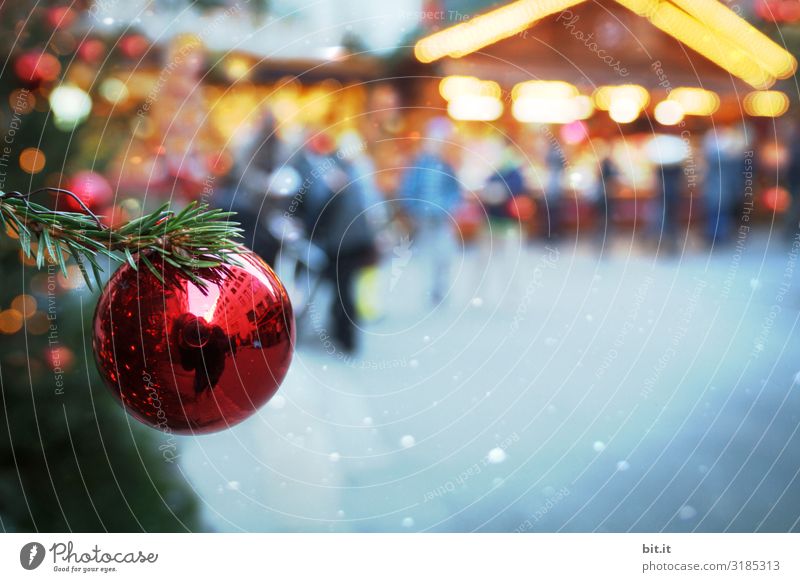 Blurred l red, shiny Christmas tree ball, hanging on fir branch in green, on illuminated Christmas market. Christmas mood with Christmas tree on Christmas market, illuminated with a lot of light, in the twilight. Evening mood on Christmas market with jewellery