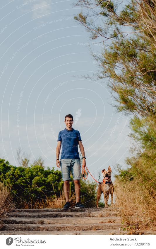 Happy adult man walking with dog along rural pathway happy casual cheerful leash brown countryside wooden tree greenery daylight pet together lifestyle smile